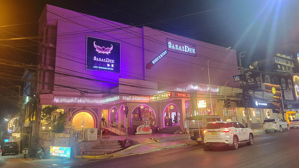 Exterior view of the Sabai Dee Body Massage building at the corner of Soi 2. The multi-story structure is brightly lit with vivid purple neon lights and displays a large 'SABAIDEE' sign alongside a mask-themed logo. The entrance area features arches and additional lighting. Cars and a baht bus are parked on the street in the foreground.