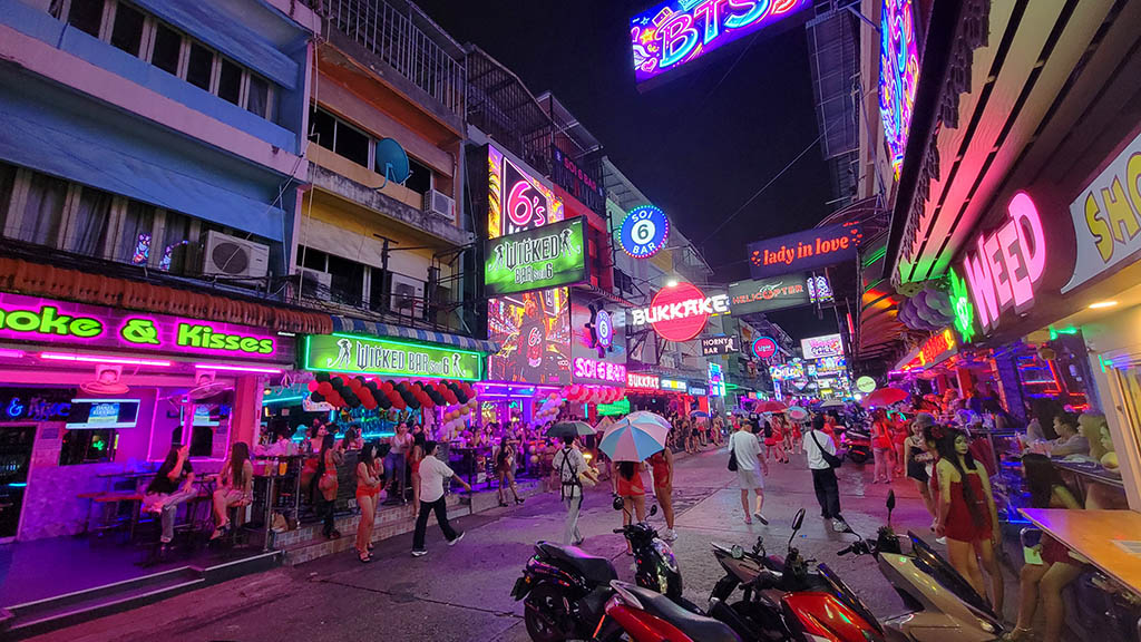 Nighttime view looking down Pattaya's vibrant Soi 6 beer bar street, bathed in glowing pink, green, and neon signage. Groups of bar hostesses in red and orange outfits stand in open-air entrances outside venues like Wicked Bar and Smoke & Kisses, with pedestrians and parked motorbikes along the street.
