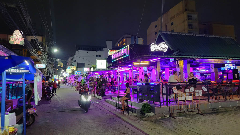 Night street view of the open-air beer bar complexes along Soi 7 in Pattaya, featuring purple neon lighting, outdoor counter seating, and the illuminated Luxor Bar sign in the background.