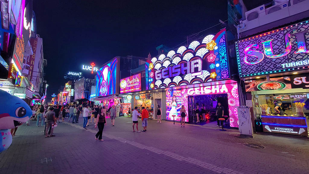 A night scene of Walking Street in Pattaya, filled with a diverse crowd of pedestrians on the paved street. The right side features brightly lit venues, including a Geisha-themed bar with a complex, multi-colored LED facade displaying fan patterns and text, and a Sultan Turkish Special Ice Cream stand. Other prominent signs include 'AURA', 'JALWA', and a large 'LUCIFER' sign. Staff members in black uniforms stand at the Geisha bar entrance.